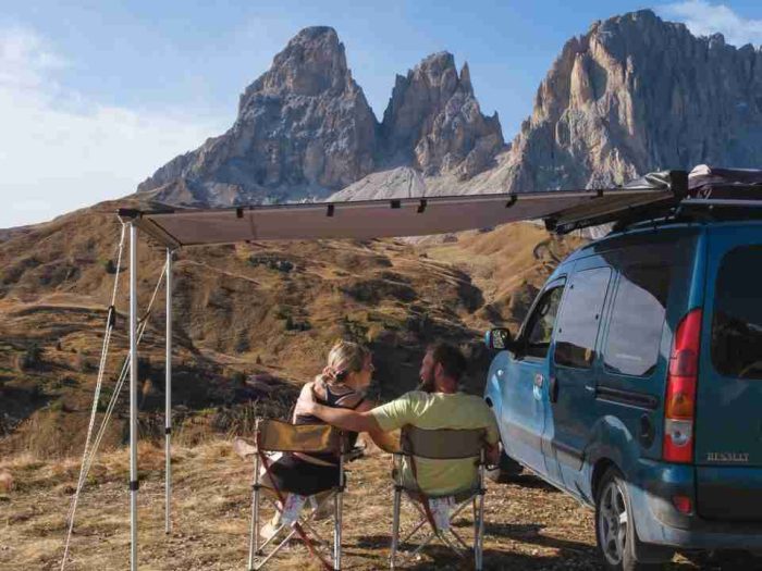 A couple in chairs hugging under a side awning on a campervan with a backdrop of mountains.